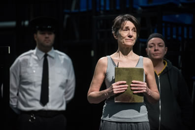 Hanna/Prospero in gray tanktop and sweatpants clutching a book, with a guard in white shirt, black tie and police hat behind her on the left, the inmate playing Calaban in black jacket and black stocking cap behind her on the right
