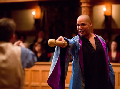 Prospero in blue and purple robe with a block Egyptian shirt open to the chest points a gold scepter at Ferdinand, whose back we see in a tan vest and blue shirt.