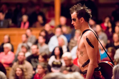 Ariel stands topless with feather tatoos on his arm and waist, starburst makeup arond his eyes, spiked hair, and a banjo draped over his should, with the Blackfriars Playhouse audience in the background.