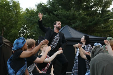 Macbeth, in black with tartan sash across his chest under his jacket, mimes holding aloft a cup as the other members of the cast, sitting around him, likewise mime toasting with cups (Lady Macbeth stands next to Macbeth) as the gray-cloaked figure of Banquo sits with his back to us to the right. In the background are large black and brown tents and trees.