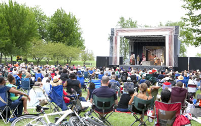 An Elizabethan costumed performance on a temporary stage in the background with people of all types sitting in chairs and on the lawn watching