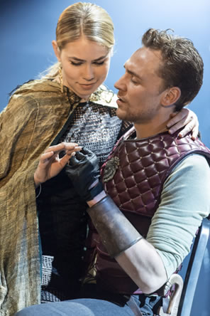 Blond and beautiful Virgilia in chain-mail patterned modern midi dress and gold cape sits on the lap of beautiful Coriolanus in green t-shirt and leather breastplate.