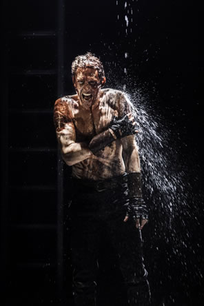 Shirtless Coriolanus, covered in blood, screams under a shower of water.