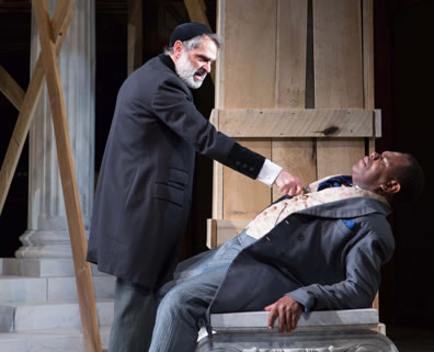 Shylock in skullcap and long black coat with gray pants presses his knife to the breast of Antoine leaning back on a platform and wearing a three-piece suit. In the background are the columns and construction scaffold