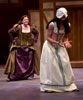 Katherina in purple Elizabethan dress with gold brocade overlay runs toward Bianca in white underdress and tan girdle, her hands bound and head covered in a cloth.