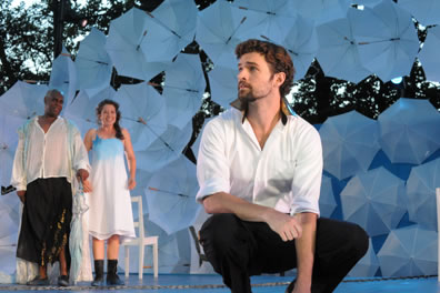 Ferdinand squatting at the front center of the picture in white shirt with gold collar and black pants, in the back ground Prospero in white shirt, ragged black pants ending at his knees, and Miranda in simple white dress with blue shading and black boots, behind them a chair, and the umbrellas of the stage wall, with real trees beyond those