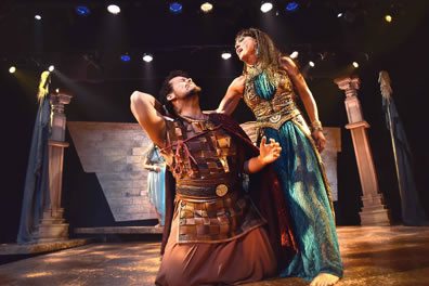 Cleopatra in a blue sheen dress with gold vest and train yanks the hair of Eros, on his knees in Roman soldiers uniform of leath vest and brown skirt. Pillars and a stone wall are in the background.