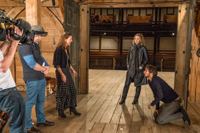 Backstage wood walls and floor, camera man to left, crewman next to him, an actress in long blue-checkered dress, blue short sweater and high-heeled boots, Cattral in long leather coat, scarf, and leather boots watching, and actor, beard, blue sweat shirt and blue jeans, kneeling on the ground; through the door you can see the stage and seating of the Globe theater