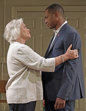Volumnia, short gray hair, white jacket blouse, gray pants, grasps Martius's arms as she looks up at him and he, in gray suit, down sheepishly at her.