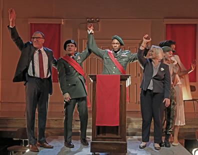 Menenius waves with right hand, wearing dark gray suit, white shirt, muted red tie, and black suspenders; Comininius in army service uniform and beret with a red sash holds up Coriolanus's right arm; Coriolanus behind podium in army service uniform, green beret, and red sash; Volumnia, holding up her son's left hand, in breay jacket, blouse, and pants; Young Martius in camoflauge pants and fatigue t-shirt waves to side; Virgilia behind her in simple tan blouse and skirt with white long coat over her shoulders, waving.
