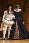 Sebastian in white Elizabethan wear--puffy pans and cap--stands next to Olivia in black dress with white ruff colar and a crown-like headband.