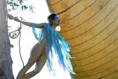 Arial, in silver leaotard, blue shreddy fabric as a shawl, and blue makeup across her forehead and down by her eyes, holds onto a rope around a real tree trunk and leans out with a ship's sail behind her