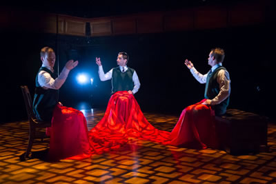 Three boys, wearing sweater vests and ties, mime sewing as they sit across from each other with a single red fabric covering their laps and legs.