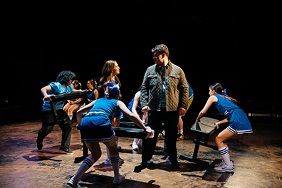 Beth wearing a blue cheerleader uniform looks up at Mac wearing an olive flack jacket over a blue football jersey and black pants, standing opposite each other as three cheerleaders and a football player carry and place benches for the next scene and two other cheerleaders sit side-by-side in the background in position to speak their first lines.