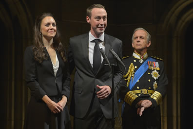 Kate in black dress suit with white shirt, hands folded at her waist stands next to William in gray suit and tie with white shirt standing at a pair of microphones. Standing slightly behind them, Charles in dress uniform with medals, blue sash, and gold braids watches with his hands clenched at his waist.