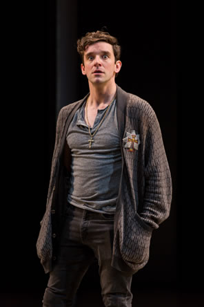 Hamlet in gray t-shirt and jeans and sweater with a cross emblem on the lapel and a cross necklace around his neck, his hands shoved in the sweater's pockets, and his eyes so wide the white surrounds the pupils.