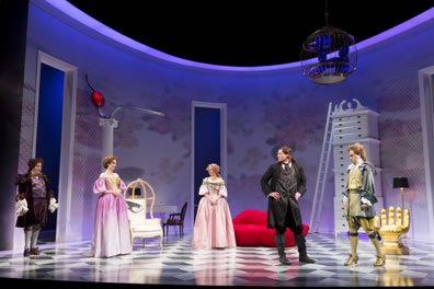 Overall photo of the set (with gold hand chair, red lipped sofa, eye-backed chair, spoon-and-cherry topped pillar, towering chest of drawers, and dachsund balloon sculpture) with members of the company: Acaste in frilly-cuffed jacket and short pants, Celimene in purple dress, Eliante in ink dress with white blouse, Frank in black suit and boots, and Philinte in shimmering green jacket, blouse, pants, and high-heeled shoes, plus blue ribbons and bows at his knees)