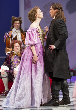 Celimene in a purple French classic dress with gold front stands face to face with Frank in black long-coat suit as two suitors in frilly, multicolored French classic courtier's suits an large, curly wigs watch in the background