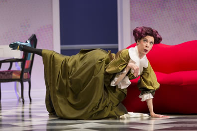 Arsinoe in a green dress and purple wig with hair curles sticking out to the side kneels on the floor, one hand up and one black-stockinged leg extended like a dog. 