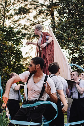 Production photo of Gower on a door carried by men, with other men twirling blue ribbons, and tall trees in the background