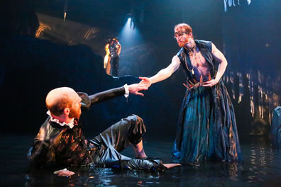 Prospero, in vest and billowing skirt, reaches his hand down to Alonso lying in the water as Miranda and Ferdinand are visible in a spotlight on the cliff at the back of the stage.