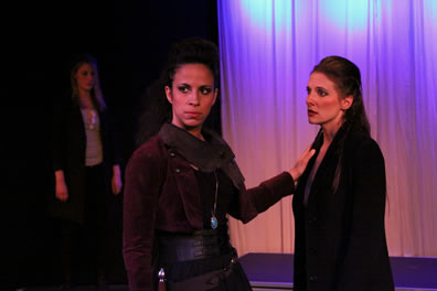 Othello, in suede jacket, thick belt, and turquoise pendant, places her hand on the shoulder of Iago, who's wearing a long, black coat. A woman in black jacket and pants and grey blouse stands in the background next to a white curtain